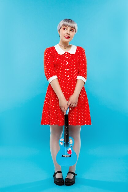 Close up portrait of beautiful dollish girl with short light violet hair wearing red dress holding ukulele over blue wall