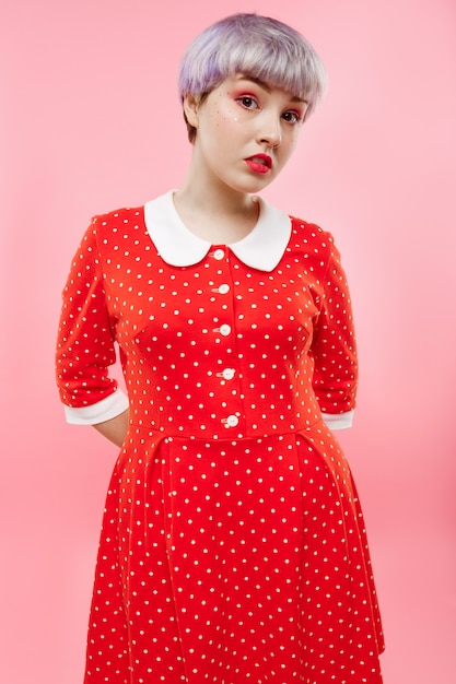 Close up portrait of beautiful dollish girl with short light violet hair wearing red dress over pink wall