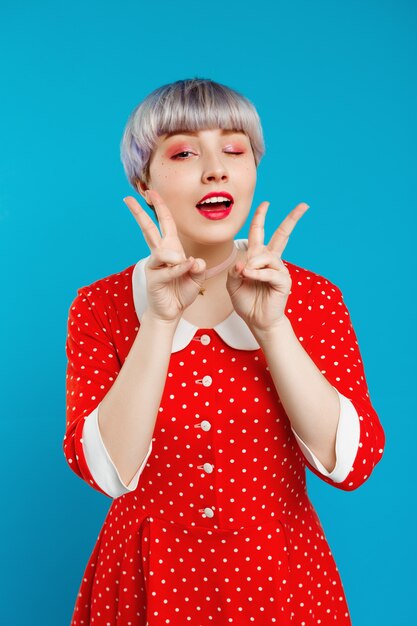 Close up portrait beautiful dollish girl with short light violet hair wearing red dresswinking showing victory gesture  over blue wall
