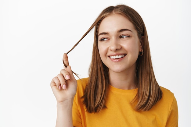 Free photo close up portrait of beautiful girl playing with hair strand, smiling and looking aside relaxed, posing in yellow t-shirt, carefree expression, white background