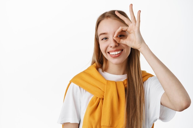 Close up portrait of beautiful happy girl with white smile showing OK okay gesture near eye approve something good pleased by good quality praise product standing over studio background