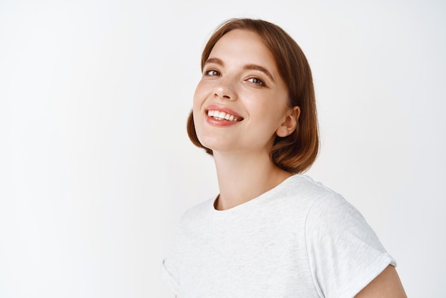 Close up portrait of beautiful lady with natural makeup smiling and looking joyful at camera standing against white background