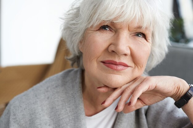 Close up portrait of beautiful positive middle aged Caucasian lady on retirement daydreaming at home, thinking about her grandchildren. Elegant gray haired grandmother spending time indoors