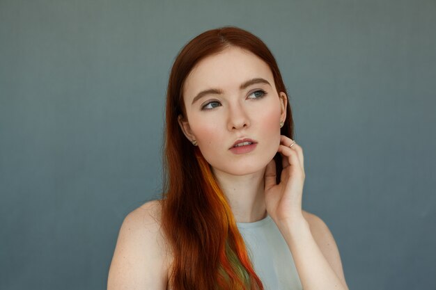 Close up portrait of a beautiful redhead female model with colored strands in hair and green eyes having thoughtful serious look, and mouth wide open. Pretty ginger girl posing on blue wall