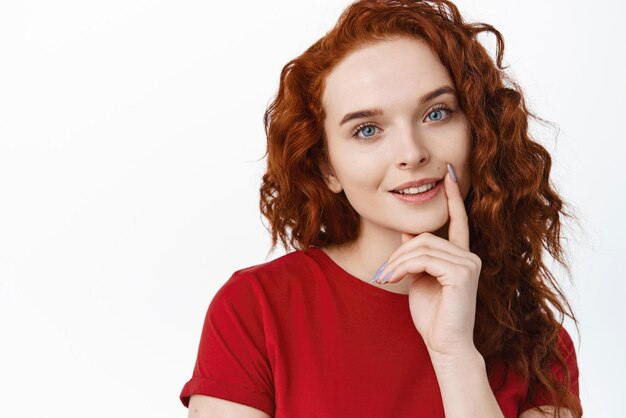 Close up portrait of beautiful redhead female model with long curly hairstyle touching lip and smiling flirty at camera look thoughtful and intrigued standing over white background