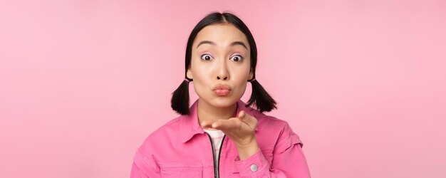 Close up portrait of beautiful silly asian girl sending air kiss mwah at camera blowing standing over pink background