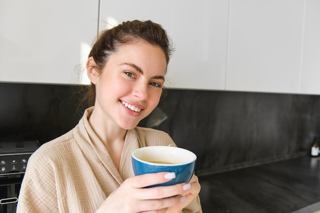 Close up portrait of beautiful smiling woman holding cup of coffee posing in the kitchen enjoying