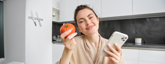 Foto gratuita ritratto ravvicinato di una bella donna sorridente che tiene pomodoro fresco seduto in cucina con lo smartphone