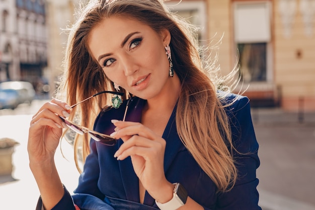 Close-up portrait of beautiful woman dressed in stylish blue jacket walking in autumn sunny street wearing elegant sunglasses and earrings