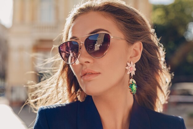 Close-up portrait of beautiful woman dressed in stylish blue jacket walking in autumn sunny street wearing elegant sunglasses and earrings