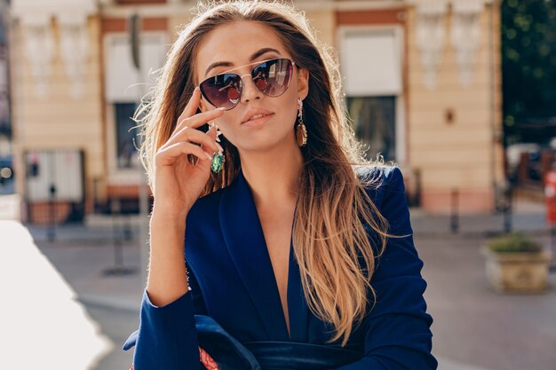 Close-up portrait of beautiful woman dressed in stylish blue jacket walking in autumn sunny street wearing elegant sunglasses