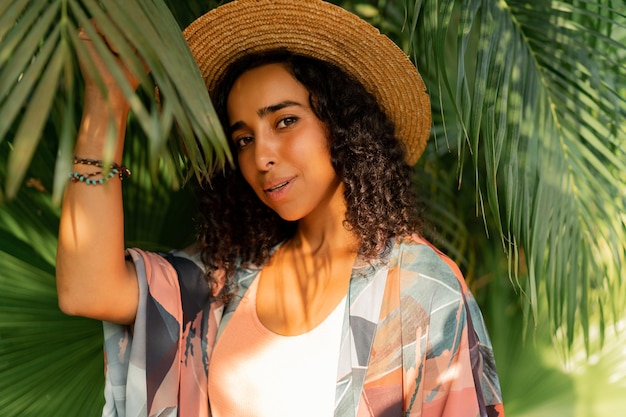Ritratto ravvicinato di bella donna con cappello di paglia e abito estivo elegante in posa nel giardino tropicale