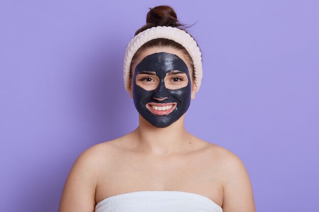 Close up portrait of beautiful woman with facial black mask isolated over lilac wall, girl with white towel on her body and bare shoulders, satisfied and happy smile, doing cleansing procedure.