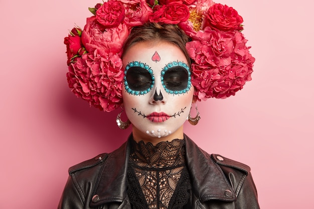 Close up portrait of beautiful woman with traditional mexican face painting, has eyes shut, wears wreath made of aromatic flowers, black attire, poses over pink wall