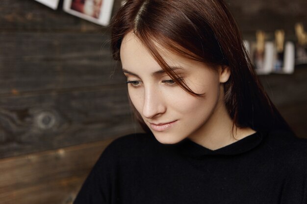 Close up portrait of beautiful young European female with chocolate hair looking down with shy smile while having rest at cozy restaurant during lunch