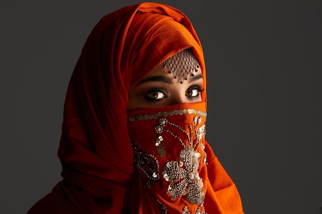 Close-up portrait of a beautiful young female with smoky eyes and jewelry on the forehead, wearing the terracotta hijab decorated with sequins. She is posing sideways and looking at the camera on a da