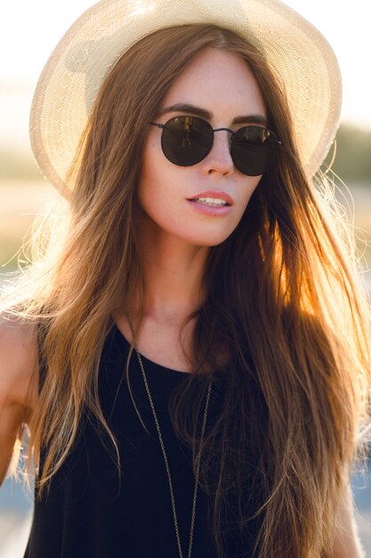 Close-up portrait of a beautiful young girl with long dark hair wearing straw hat, and dark sunglasses. She smiles slightly