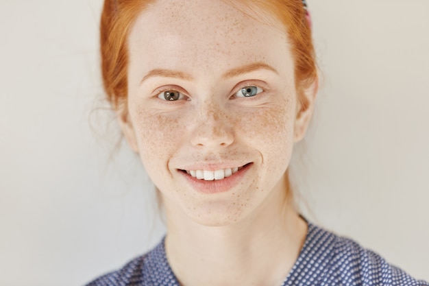 Free photo close up portrait of beautiful young redhead model with different colored eyes and healthy clean skin with freckles smiling joyfully, showing her white teeth, posing indoors. heterochromia in human