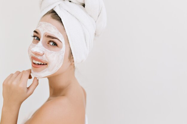 入浴後タオルで美しい若い女性のクローズアップの肖像画は、彼女の顔に化粧品のマスクを作ります。