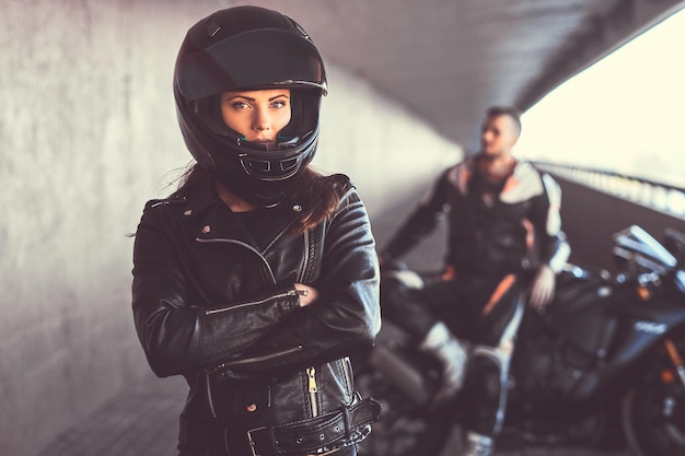 Close-up portrait of a biker girl wearing a leather jacket and helmet with her arms crossed next to her superbike inside the bridge.