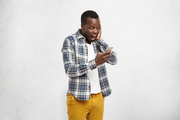 Free photo close up portrait of black shocked hipster in stylish and trendy yellow pants, holding smartphone in one hand touching his head