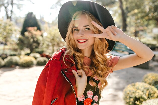 Close-up portrait of blinde woman in black wide-brimmed hat and red jacket showing peace sign in park.