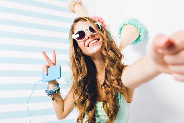 Close-up portrait of blissful girl in sunglasses and trendy bracelets posing with peace sign. Charming young woman with long hair making selfie holding phone and listening favorite song.