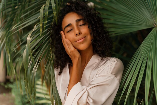 Close up portrait of  blissful  woman with wavy hairs posig  in tropical garden. Wearing elegant silk robe. Spa and  skincare  concept.