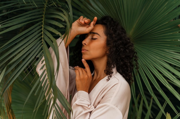 Close up portrait of  blissful  woman with wavy hairs posig  in tropical garden. Wearing elegant silk robe. Spa and  skincare  concept.