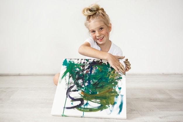 Close-up portrait of blond European little girl with hair bun and freckles smiling with all her teeth. Holding on her knees picture that she painted for her parents, feeling proud of herself. People a