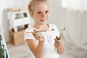 Free photo close-up portrait of blonde cute european little girl with paint on her freckled face and hair bun smiling with all her teeth holding a bunch of brushes in her hands. cheerful girl messed up her white