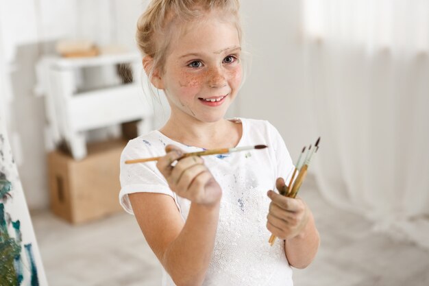 Close-up portrait of blonde cute European little girl with paint on her freckled face and hair bun smiling with all her teeth holding a bunch of brushes in her hands. Cheerful girl messed up her white