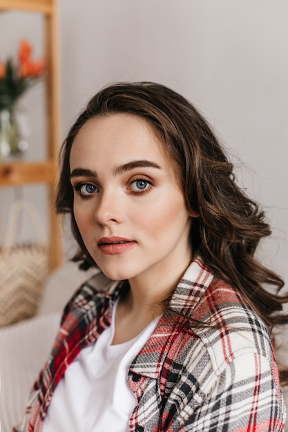 Close-up portrait of blue-eyed brunette curly young woman in plaid shirt and white tee looking into front and sitting on beige sofa in living room