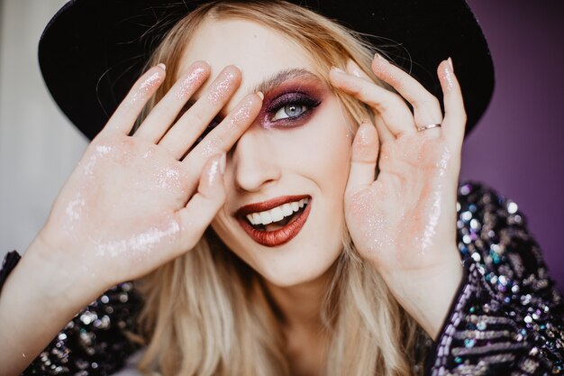 Close-up portrait of blue-eyed good-humoured woman in black hat. Caucasian lovable girl with dark makeup smiling.