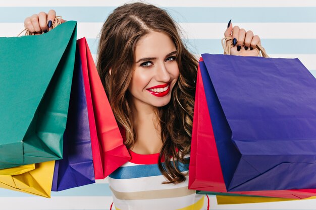 Close-up portrait of blue-eyed woman with black nails holding colorful packages. Indoor photo of blissful dark-haired girl smiling