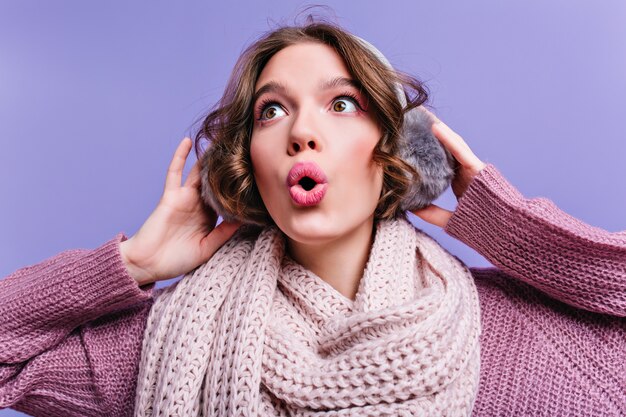 Close-up portrait of brown-eyed short-haired girl touching fur headphones. surprised european lady in woolen scarf isolated on purple wall.