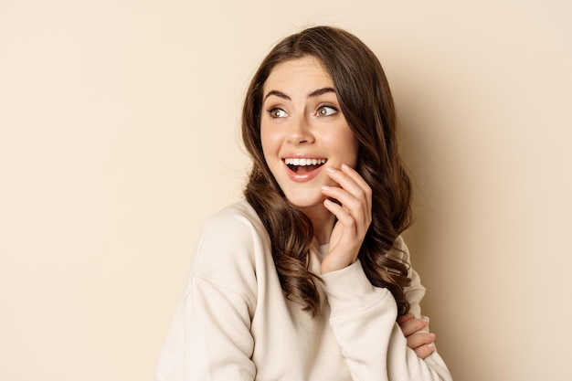 Close up portrait of brunette feminine woman turn back, look behind with surprised face expression, standing against beige background.