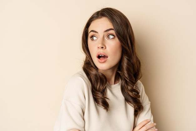 Close up portrait of brunette feminine woman turn back, look behind with surprised face expression, standing against beige background