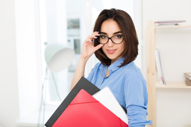 Close-up portrait of a brunette girl standing in office. She is holding folders in one hand and glasses on face in another.