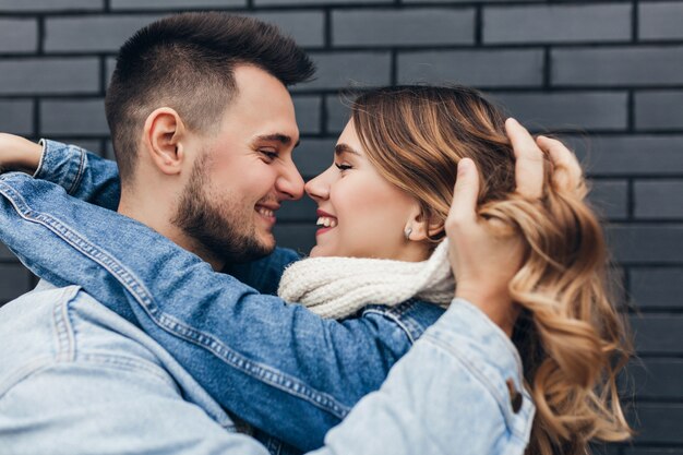 Close-up portrait of brunette guy playing with girlfriend's hair. Blithesome european lady looking into husband's eyes on bricked wall.