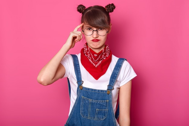 Close up portrait of brunette student, wears round eyewear and red bandana on neck, curves lips, keeps fore finger on temple