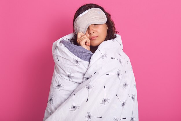 Close up portrait of brunette woman peeping from sleeping mask, does not want to wake up, keeps eyes closed, wearing white blanket