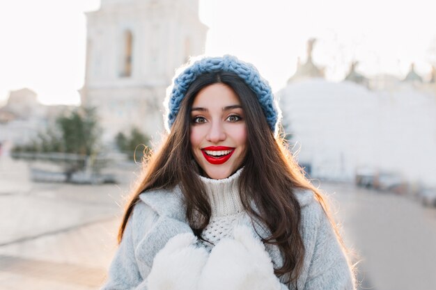 Крупным планом портрет брюнетка женщины с красными губами, улыбаясь на размытом городе. Наружное фото беззаботной девушки в голубой вязаной шапке и теплых перчатках, позирующих с удивленным выражением лица.