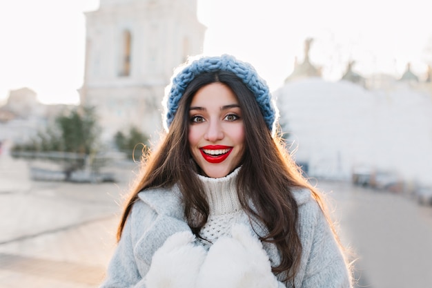 Крупным планом портрет брюнетка женщины с красными губами, улыбаясь на размытом городе. Наружное фото беззаботной девушки в голубой вязаной шапке и теплых перчатках, позирующих с удивленным выражением лица.