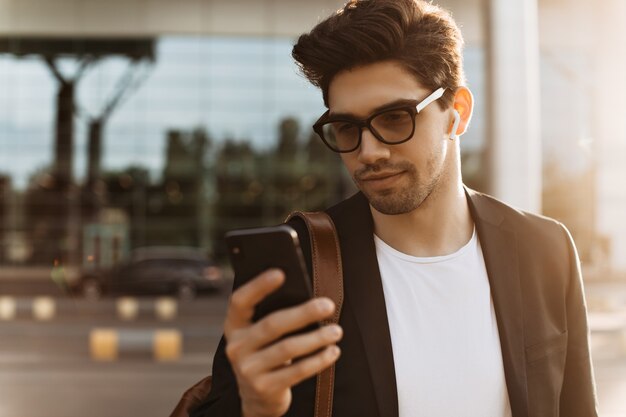 Close-up portrait of brunette young guy in eyeglasses, white t-shirt and black jacket holding phone and messaging