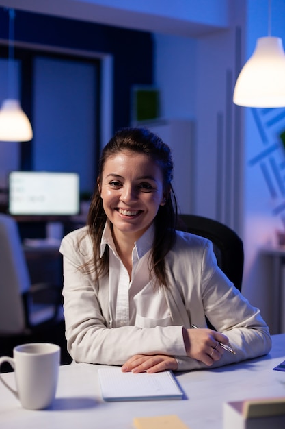 Close up portrait of business woman smiling at camera after drinking cup of coffee sitting at desk in business office late at night