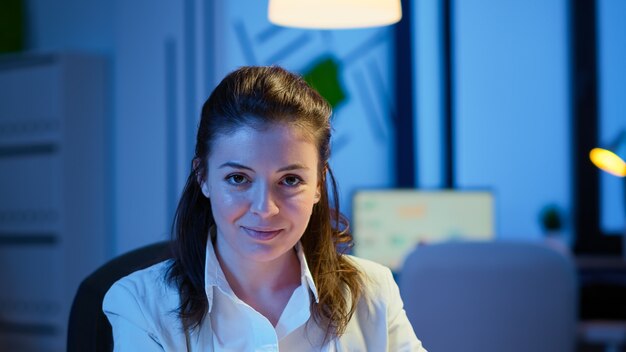 Close up portrait of business woman smiling at camera after reading mails on laptop sitting at desk in start-up company late at night. Focused employee using technology network wireless doing overtime