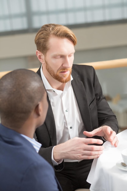 Close-up portrait of businessman explaining something to his foreign business partner while sitting at table during business meeting in restaurant.