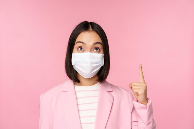 Close up portrait of businesswoman asian lady in office suit and medical face mask looking and pointing up showing company logo or banner on top pink background