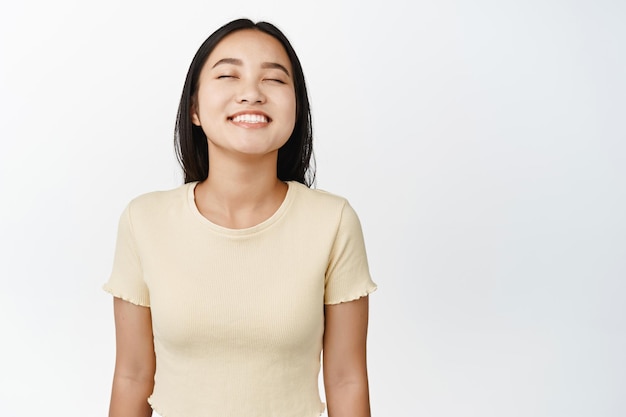 白い背景の上に黄色のTシャツに立っている何かを空想にふける目を閉じて笑っているのんきなアジアの女性の肖像画をクローズアップ